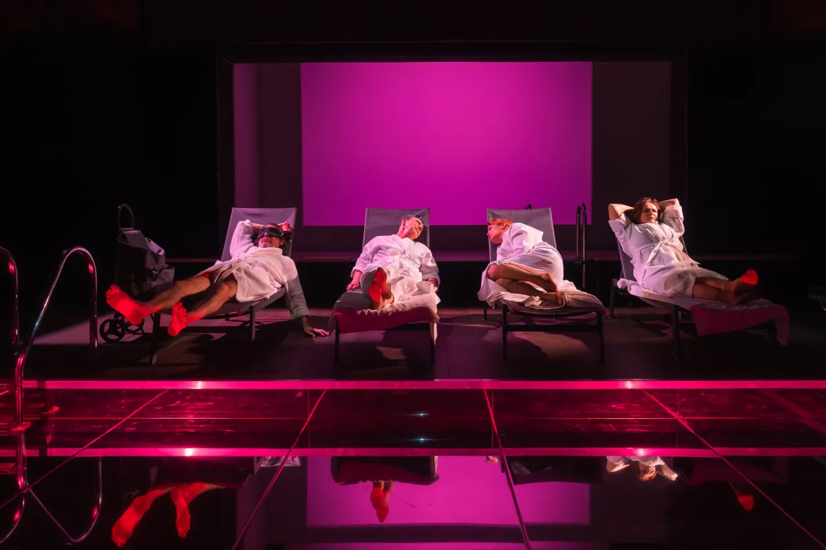 Scena ze spektaklu „Pływalnia” w Teatrze Jaracza. Czworo aktorów w białych szlafrokach leży obok siebie na leżakach basenowych ustawionych w głębi sceny. Całość tonie w intensywnym, różowo-fioletowym świetle reflektorów. Na pierwszym planie znajduje się gładka, ciemna podłoga, w której odbijają się postacie i zarysy leżaków, a po lewej stronie widać metalową drabinkę basenową.