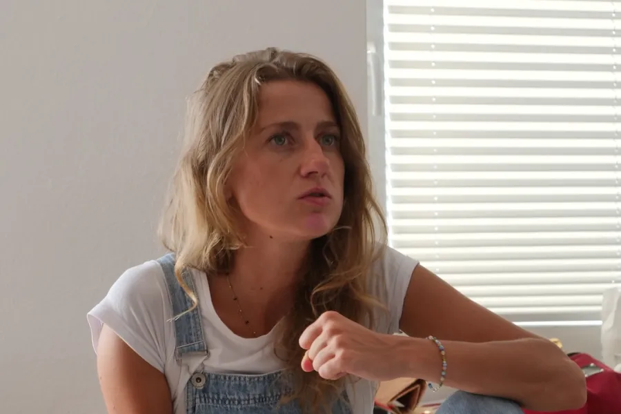 Zdjęcie przedstawia próbę stolikową do spektaklu „Ifigenia ze Splott”. Przy stole ze zgiętym kolanem, położonym na nim łokciu i zaciśniętą pięścią siedzi przy stole Agnieszka Skrzypczak. Aktorka patrzy przed siebie a kąciki jej ust są lekko opuszczone przy równoczesnym ich otwarciu. Przed aktorką na stole leży scenariusz, telefon, kubek w kwiaty, fotografie i długopis.  