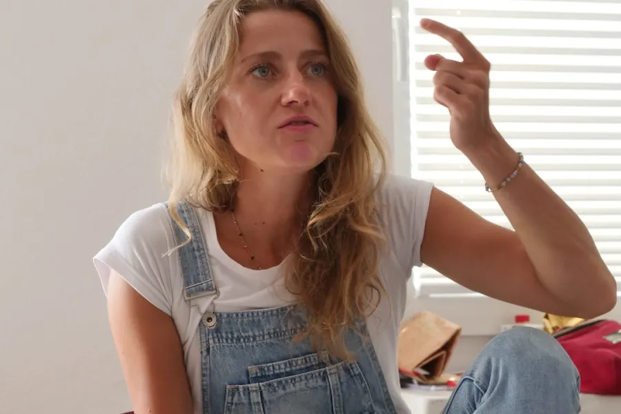 Zdjęcie przedstawia próbę stolikową do spektaklu „Ifigenia ze Splott”. Przy stole ze zgiętym kolanem i lekko podniesionym łokciem siedzi przy stole Agnieszka Skrzypczak. Wskazujący, zgięty palec aktorki jest lekko skierowany ku górze. Wzrok Agnieszki Skrzypczak skierowany jest przed siebie a kąciki jej ust są bardziej opuszczone w dół. Przed aktorką na stole z zielonym obrusem znajduje się scenariusz, fotografie, telefon, kubek w kwiaty i długopis.