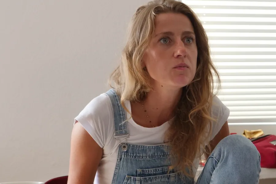 Zdjęcie przedstawia próbę stolikową do spektaklu „Ifigenia ze Splott”. Przy stole z lekko pochyloną do przodu, zgarbioną postawą i zgiętym kolanem siedzi Agnieszka Skrzypczak. Oczy aktorki są szeroko otwarte a kąciki jej ust lekko ściągnięte do środka. Na stole przed aktorką leży scenariusz, kubek w kwiaty, fotografie i telefon.