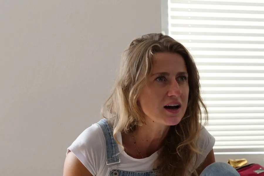 Zdjęcie przedstawia próbę stolikową do spektaklu „Ifigenia ze Splott”. Przy stole z lekko pochyloną do przodu, zgarbioną postawą i zgiętym kolanem siedzi Agnieszka Skrzypczak. Wzrok aktorki jest skierowany przed siebie, usta są otwarte a ręce zwisają wzdłuż ciała. Na stole przed aktorką leży scenariusz, kubek w kwiaty, fotografie i telefon.