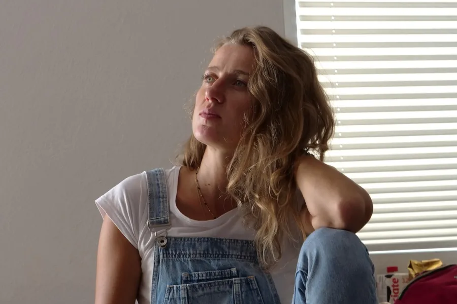 Zdjęcie przedstawia próbę stolikową do spektaklu „Ifigenia ze Splott”. Przy stole ze zgiętym kolanem i opartym na nim łokciu siedzi Agnieszka Skrzypczak. Ręka oparta na kolanie jest ugięta i sięga tyłu głowy aktorki a druga ręka zwisa wzdłuż ciała. Głowa Agnieszki Skrzypczyk jest zwrócona w prawą stronę i lekko pochylona do tyłu. W tę samą stronę, również sięga wzrok aktorki. Na stole przed aktorką leży scenariusz, kubek w kwiaty, fotografie i telefon.