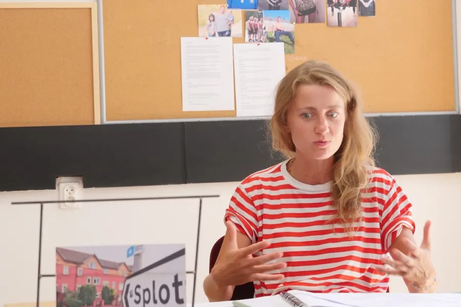  Zdjęcie przedstawia próbę stolikową do spektaklu „Ifigenia ze Splott”. Przy stole siedzi skupiona Agnieszka Skrzypczak z dwoma zgiętymi rękoma. Palce dłoni aktorki są mocno od siebie rozstawione a same dłonie wewnętrzną stroną do siebie lekko wyciągnięte do przodu. Oczy Agnieszki Skrzypczak zwrócone są poniżej linii obiektywu a kąciki jej ust mocno ściągnięte do siebie. Na stole oprócz zeszytu z ołówkiem znajduje się zdjęcie przedstawiające brytyjską kamienicę i znak z nazwą dzielnicy Splott.