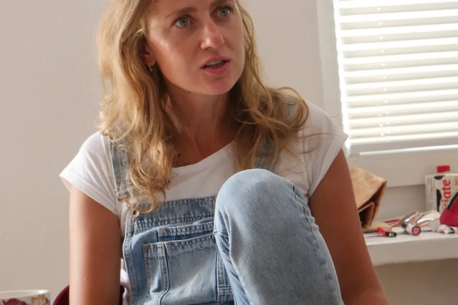 Zdjęcie przedstawia próbę stolikową do spektaklu „Ifigenia ze Splott”. Przy stole ze zgiętym kolanem i lekko podniesionymi łokciami siedzi przy stole Agnieszka Skrzypczak. Wzrok Agnieszki Skrzypczak skierowany jest przed siebie a usta lekko otwarte. Przed aktorką na stole z zielonym obrusem znajduje się scenariusz, fotografie, telefon, kubek w kwiaty i długopis.