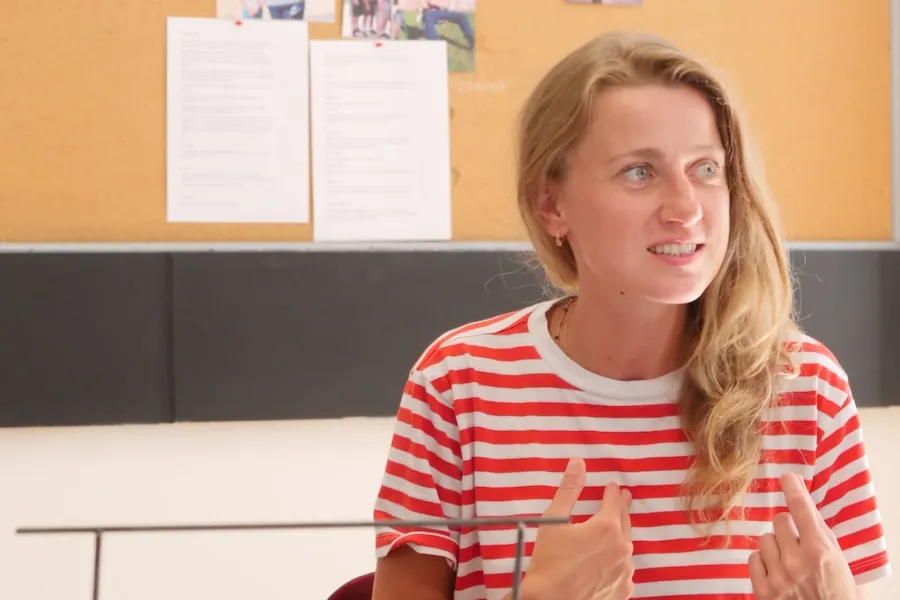 Zdjęcie przedstawia próbę stolikową do spektaklu „Ifigenia ze Splott”. Przy stole siedzi Agnieszka Skrzypczak ze zgiętymi w łokciach rękami skierowanymi na siebie. Wzrok aktorki jest skierowany poza obiektyw, a jej zęby są lekko zaciśnięte. Przed aktorką na stole znajdują się fotografie i makieta osiedlowego trzepaka. Na makiecie jest oparte zdjęcie angielskiej kamienicy z znakiem „Splott”. Oprócz trzepaka na stole znajduje się telefon, scenariusz i długopis. Z tyłu wisi na ścianie tablica korkowa.