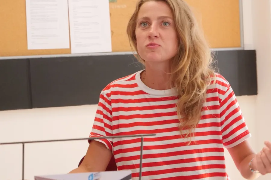 Zdjęcie przedstawia próbę stolikową do spektaklu „Ifigenia ze Splott”. Przy stole siedzi Agnieszka Skrzypczak z rozłożonymi rękami. Wzrok aktorki jest skierowany ponad obiektyw, a jej wargi są lekko wysunięte do przodu. Przed aktorką na stole znajdują się fotografie i makieta osiedlowego trzepaka. Na makiecie jest oparte zdjęcie angielskiej kamienicy z znakiem „Splott”. Oprócz trzepaka na stole znajduje się telefon, scenariusz i długopis. Z tyłu wisi na ścianie tablica korkowa. 