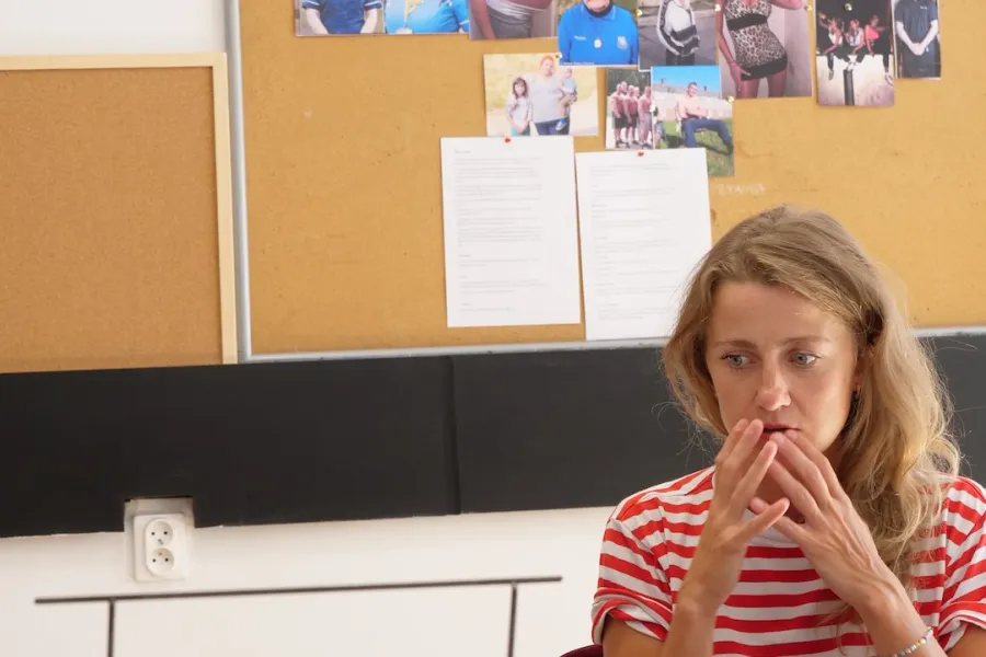 Zdjęcie przedstawia próbę stolikową do spektaklu „Ifigenia ze Splott”. Przy stole siedzi Agnieszka Skrzypczak ze złożonymi przy twarzy dłońmi. Wzrok aktorki jest skierowany poniżej obiektywu. Przed aktorką na stole znajdują się fotografie i makieta osiedlowego trzepaka. Na makiecie jest oparte zdjęcie angielskiej kamienicy z znakiem „Splott”. Oprócz trzepaka na stole znajduje się scenariusz i długopis. Z tyłu wisi na ścianie tablica korkowa ze zdjęciami.  