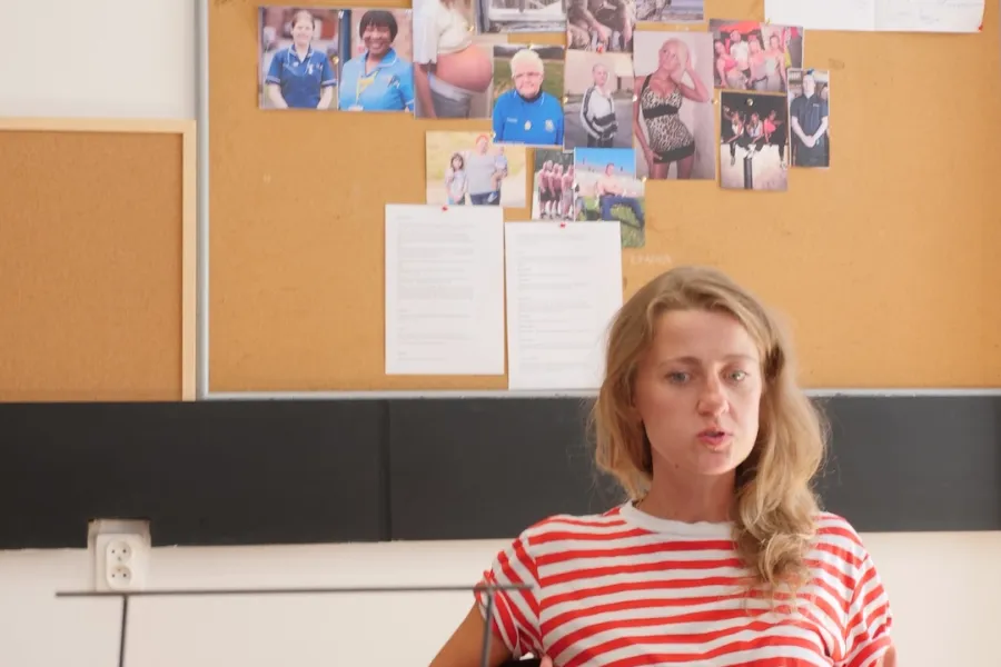 Zdjęcie przedstawia próbę stolikową do spektaklu „Ifigenia ze Splott”. Przy stole siedzi skupiona Agnieszka Skrzypczak z dwoma zgiętymi rękoma opartymi na oparciu krzesła. Palce dłoni aktorki są mocno od siebie rozstawione. Oczy Agnieszki Skrzypczak zwrócone są poniżej linii obiektywu a kąciki jej ust mocno ściągnięte do siebie. Na stole oprócz zeszytu z ołówkiem znajduje się zdjęcie przedstawiające z tyłu brytyjską kamienicę, a z przodu znak z nazwą dzielnicy Splott. 