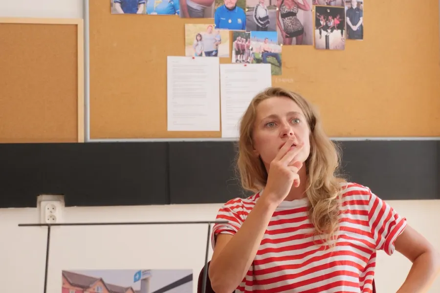 Zdjęcie przedstawia próbę stolikową do spektaklu „Ifigenia ze Splott”. Przy stole siedzi skupiona Agnieszka Skrzypczak z dwoma palcami na ustach i ze zgiętą ręką położoną na biodrze. Na stole oprócz zeszytu z ołówkiem znajduje się zdjęcie przedstawiające z tyłu brytyjską kamienicę, a z przodu znak z nazwą dzielnicy Splott. Z tyłu znajduje się korkowa tablica z wieloma zdjęciami różnych ludzi, tekstami oraz napisem Cardiff.