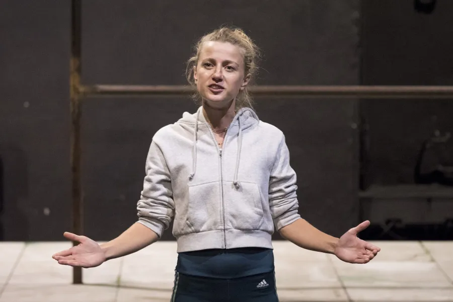 Zdjęcie przedstawia drugą próbę do spektaklu „Ifigenia ze Splott”. Agnieszka Skrzypczak stoi w centrum fotografii. Fotografia jest przybliżona, aktorkę widać od pasa w górę. Stoi na wprost obiektywu. Głowę ma skierowaną minimalnie w prawo, w tą stronę też patrzy. Ręce ma podniesione na wysokość bioder, dłonie otwarte, skierowane na zewnątrz. Aktorka ma otwarte usta. Za aktorką widać fragment lewej strony scenografii trzepaka osiedlowego na podeście.