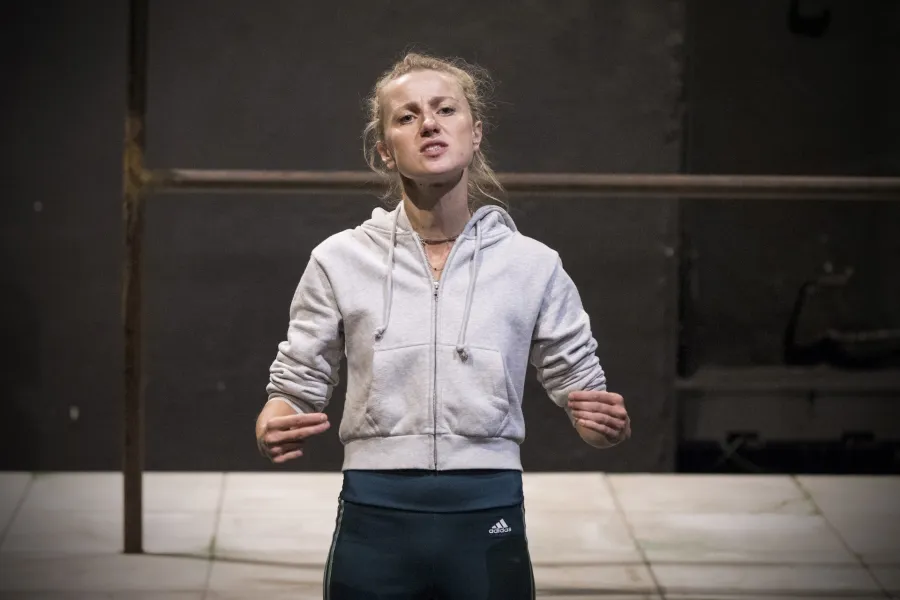 Zdjęcie przedstawia drugą próbę do spektaklu „Ifigenia ze Splott”. Agnieszka Skrzypczak stoi w centrum fotografii. Fotografia jest przybliżona, aktorkę widać od pasa w górę. Stoi na wprost obiektywu. Ręce ma podniesione na wysokość bioder. Wskazuje na siebie palcem wskazującym i środkowym prawej ręki, oraz lewą dłonią. Głowę ma lekko podniesioną, patrzy się w obiektyw. Usta ma nieznacznie otworzone. Za aktorką widać fragment lewej strony scenografii trzepaka osiedlowego na podeście. 