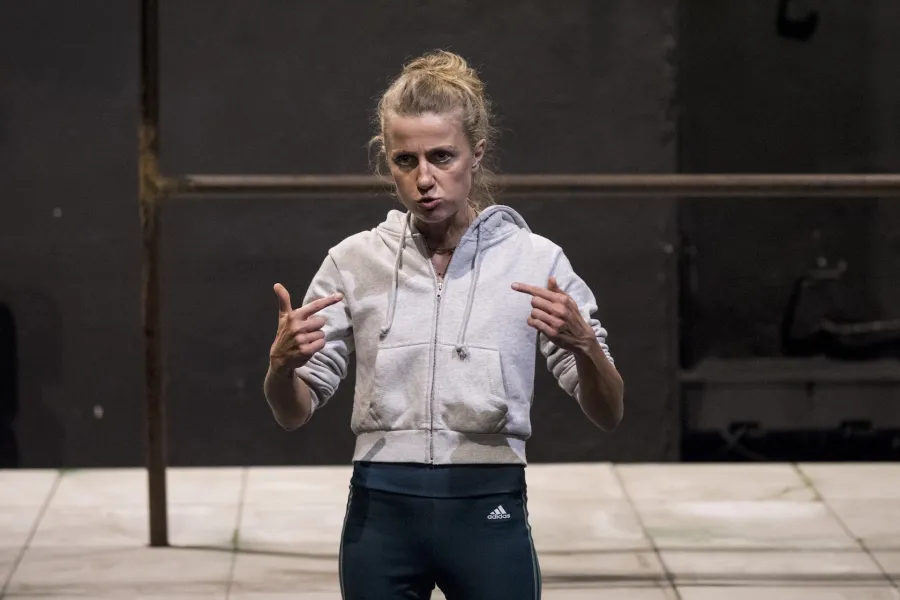 Zdjęcie przedstawia drugą próbę do spektaklu „Ifigenia ze Splott”. Agnieszka Skrzypczak stoi w centrum fotografii. Fotografia jest przybliżona, aktorkę widać od pasa w górę. Stoi na wprost obiektywu. Ręce ma nad biodrami. Wskazuje na siebie palcami wskazującymi obydwu rąk. Głowę ma skierowaną w prawo, w tą stronę też patrzy. Usta ma nieznacznie otworzone. Za aktorką widać fragment lewej strony scenografii trzepaka osiedlowego na podeście. 