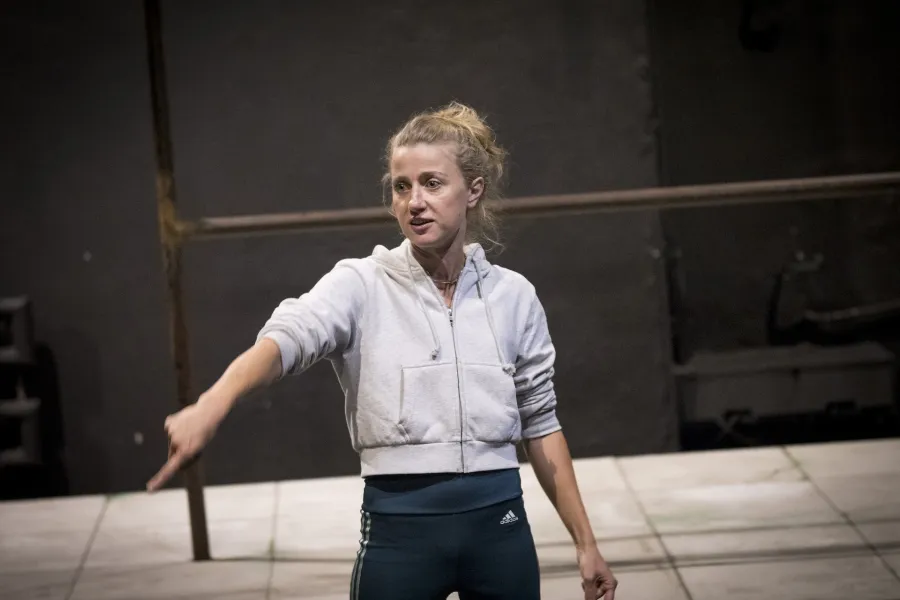 Zdjęcie przedstawia drugą próbę do spektaklu „Ifigenia ze Splott”. Agnieszka Skrzypczak stoi w centrum fotografii. Fotografia jest przybliżona, aktorkę widać od pasa w górę. Biodro aktorki jest lekko skręcone do boku. Aktorka ma głowę odwróconą w prawą stronę. Łokieć prawej ręki ma na wysokości klatki piersiowej, palcem wskazującym wskazuje lewy dolny róg fotografii. Wzrok aktorki jest skierowany w tą samą stronę co palec. Usta ma lekko otwarte. Za aktorką widać fragment lewej strony scenografii trzepaka.