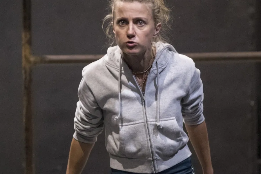 Zdjęcie przedstawia drugą próbę do spektaklu „Ifigenia ze Splott”. Agnieszka Skrzypczak stoi w centrum fotografii. Aktorka jest lekko pochylona w stronę obiektywu. Ręce ma nieznacznie za biodrami, dłonie ma otwarte. Nogi ma rozstawione. Aktorka patrzy się w obiektyw, oczy ma szeroko otwarte. Usta ma zamknięte, szczękę zaciśniętą. Za aktorką widać fragment lewej strony scenografii trzepaka osiedlowego na podeście.