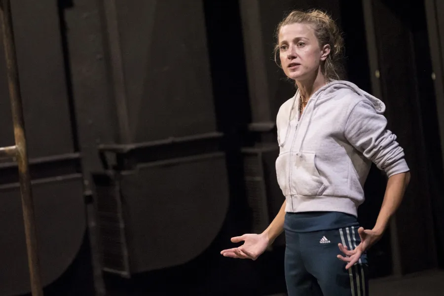 Zdjęcie przedstawia drugą próbę do spektaklu „Ifigenia ze Splott”. Agnieszka Skrzypczak stoi bokiem po prawej stronie fotografii. Aktorka patrzy w kierunku lewego końca fotografii. Ręce ma lekko zgięte. Dłonie ma otwarte, skierowane wewnętrzną stroną do góry. Usta ma lekko otwarte. Po lewej stronie fotografii widać fragment pręta scenografii trzepaka osiedlowego.