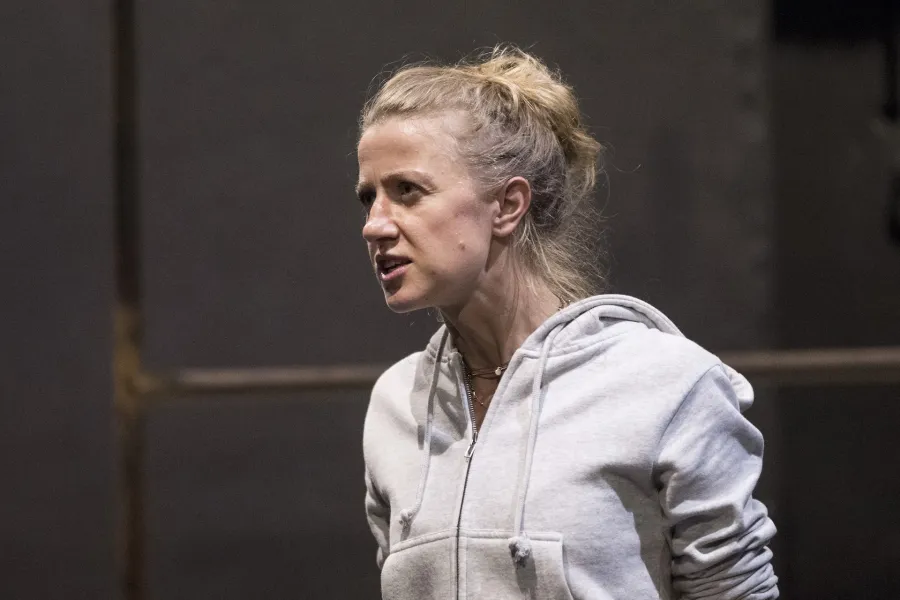 Zdjęcie przedstawia drugą próbę do spektaklu „Ifigenia ze Splott”. Agnieszka Skrzypczak stoi bokiem w centrum fotografii. Fotografia jest przybliżona, aktorkę widać od klatki piersiowej w górę. Aktorka patrzy w kierunku lewego końca fotografii. Usta ma otwarte, brwi ma zmarszczone. W tle widać fragment scenografii trzepaka osiedlowego.