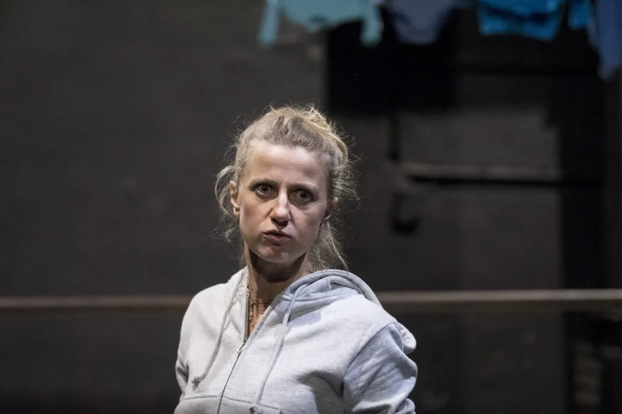 Zdjęcie przedstawia drugą próbę do spektaklu „Ifigenia ze Splott”. Agnieszka Skrzypczak stoi bokiem w centrum fotografii. Fotografia jest przybliżona, aktorkę widać od klatki piersiowej w górę. Głowę ma odwróconą w kierunku obiektywu. Oczy ma szeroko otwarte, patrzy się w obiektyw. Usta ma zamknięte. W tle widać fragment scenografii trzepaka osiedlowego. W prawym górnym rogu fotografii widać cztery sztuki błękitnej odzieży zawieszonej na górnej poprzeczce trzepaka.