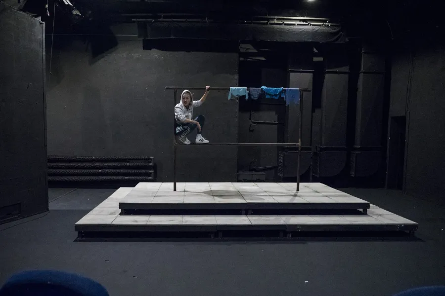 Zdjęcie przedstawia drugą próbę do spektaklu „Ifigenia ze Splott”. W centrum fotografii znajduje się scenografia trzepaka osiedlowego. Agnieszka Skrzypczak kuca na dolnej poprzeczce z lewej strony, opierając się plecami o lewy pręt. Lewą ręką trzyma górną poprzeczkę trzepaka, a prawą ma opartą na prawym kolanie. Jest lekko odwrócona w prawą stronę, nie patrzy w obiektyw. Na głowie ma założony kaptur białej bluzy. Na górnej poprzeczce trzepaka, z prawej strony, wiszą cztery sztuki błękitnej odzieży.