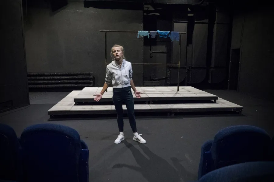 Zdjęcie przedstawia drugą próbę do spektaklu „Ifigenia ze Splott”. Agnieszka Skrzypczak stoi w centrum zdjęcia. Aktorka stoi na wprost do obiektywu, jest lekko pochylona do boku. Ręce ma lekko zgięte. Dłonie ma otwarte, skierowane wewnętrzną stroną do góry. Aktorka patrzy w obiektyw, głowę ma lekko podniesioną do góry. Ma otwarte usta. W dolnych rogach zdjęcia widać niebieskie fotele. Za aktorką widać scenografię trzepaka. Na górnej poprzeczce wisi błękitna odzież.