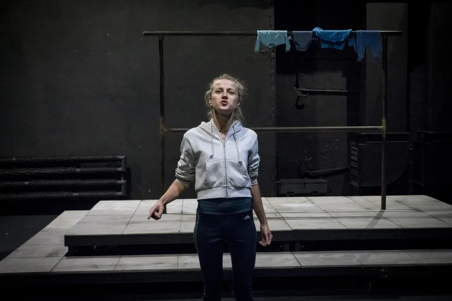 Zdjęcie przedstawia drugą próbę do spektaklu „Ifigenia ze Splott”. Agnieszka Skrzypczak stoi w centrum zdjęcia, przodem do obiektywu. Prawą rękę ma zgiętą, wskazuje palcem wskazującym w lewy dolny róg zdjęcia. Głowę ma lekko podniesioną do góry. Wzrok ma powyżej linii obiektywu. Usta ma lekko otwarte. Za aktorką widać scenografię trzepaka osiedlowego na podeście. Na górnej poprzeczce, po prawej stronie fotografii, wisi błękitna odzież.