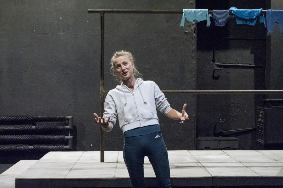 Zdjęcie przedstawia drugą próbę do spektaklu „Ifigenia ze Splott”. Agnieszka Skrzypczak stoi w centrum zdjęcia, przodem do obiektywu. Ciężar ciała aktorki skoncentrowany jest na prawej nodze. Aktorka jest lekko pochylona do tyłu. Ręce ma zgięte w łokciach. Dłonie ma otwarte, skierowane na zewnątrz. Wzrok ma skierowany w stronę prawego górnego rogu fotografii. Aktorka jest uśmiechnięta. Za aktorką widać scenografię trzepaka na podeście. Na górnej poprzeczce, po prawej stronie fotografii, wisi błękitna odzież