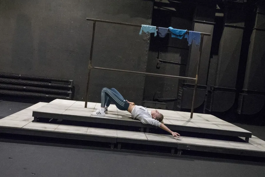 Zdjęcie przedstawia drugą próbę do spektaklu „Ifigenia ze Splott”. Agnieszka Skrzypczak znajduje się w centrum fotografii. Aktorka leży na podeście, lewym profilem do obiektywu. Ma podniesione biodra. Lewą rękę ma wyciągniętą za głowę, a prawą położoną na brzuchu. Za aktorką widać scenografię trzepaka osiedlowego. Na górnej poprzeczce, po prawej stronie fotografii, wisi błękitna odzież.