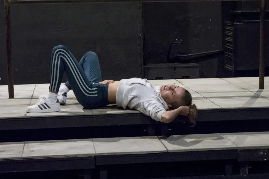 Zdjęcie przedstawia drugą próbę do spektaklu „Ifigenia ze Splott”. Agnieszka Skrzypczak znajduje się w centrum fotografii. Aktorka leży na podeście, lewym profilem do obiektywu. Aktorka ma głowę skierowaną w stronę obiektywu. Lewą ręką trzyma się za włosy, a prawą ma położoną na brzuchu. Nogi ma zgięte w kolanach. Aktorka jest uśmiechnięta. Za aktorką widać fragment scenografii osiedlowego trzepaka.