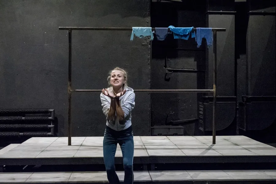 Zdjęcie przedstawia drugą próbę do spektaklu „Ifigenia ze Splott”. Agnieszka Skrzypczak znajduje się w centrum fotografii. Aktorka stoi na wprost obiektywu, jest pochylona do przodu, ma ugięte kolana. Ręce ma przed klatką piersiową, stykają się ze sobą. Dłonie są otwarte, skierowane wewnętrzną częścią w górę. Aktorka jest uśmiechnięta. Głowę ma podniesioną, wzrok skierowany ma w stronę lewego górnego rogu fotografii. Za aktorką widać scenografię trzepaka. Na górnej poprzeczce trzepaka wisi błękitna odzież.