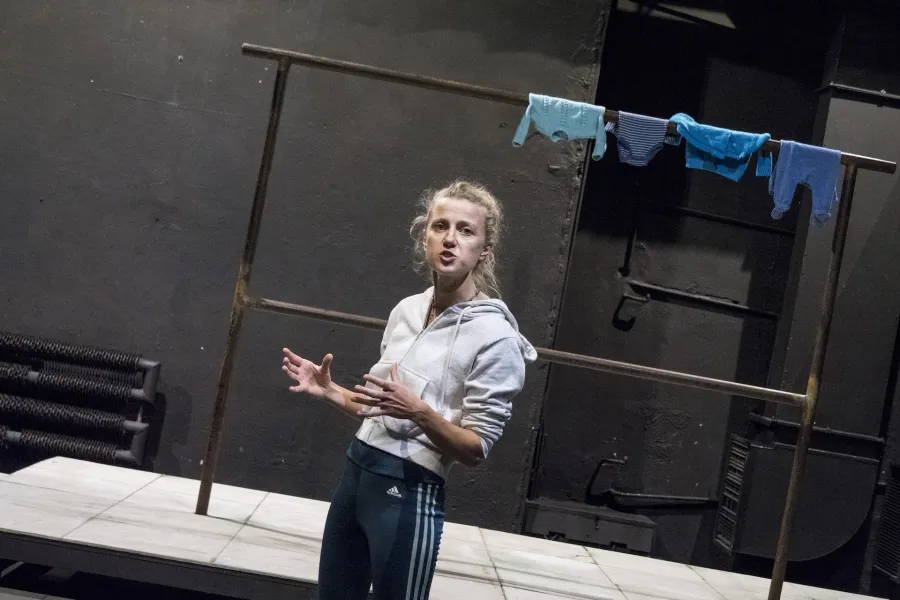 Zdjęcie przedstawia drugą próbę do spektaklu „Ifigenia ze Splott”. Agnieszka Skrzypczak znajduje się w centrum fotografii, stoi bokiem. Fotografia jest zrobiona pod kątem. Aktorka patrzy w obiektyw. Ręce ma zgięte pod kątem 90 stopni. Dłonie ma otwarte, skierowane wewnętrzną stroną do siebie. Usta ma lekko otwarte. Za aktorką widać scenografię trzepaka. Na górnej poprzeczce trzepaka wisi błękitna odzież.