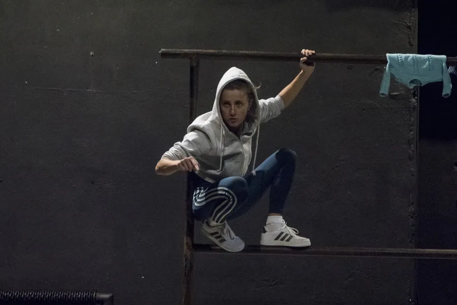 Zdjęcie przedstawia drugą próbę do spektaklu „Ifigenia ze Splott”. Agnieszka Skrzypczak kuca na scenografii trzepaka osiedlowego z lewej strony, opierając się plecami o lewy pręt. Lewą ręką trzyma górną poprzeczkę trzepaka, a prawą wskazuje w stronę kamery. Jest lekko odwrócona w prawą stronę, nie patrzy w obiektyw. Na głowie ma założony kaptur białej bluzy. W prawym rogu fotografii, na górnej poprzeczce widać zawieszoną błękitną bluzkę.