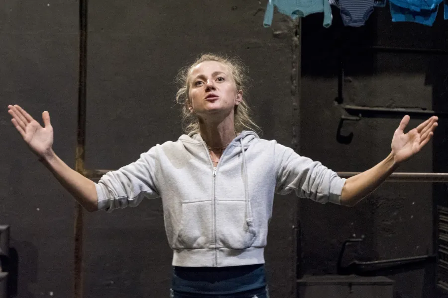 Zdjęcie przedstawia drugą próbę do spektaklu „Ifigenia ze Splott”. Agnieszka Skrzypczak znajduje się w centrum fotografii. Fotografia jest przybliżona, aktorkę widać od pasa w górę. Aktorka ma podniesioną głowę, patrzy ponad linię obiektywu. Ręce ma podniesione, szeroko otwarte, zgięte w łokciach. Dłonie ma otwarte wewnętrzną stroną w górę. Usta ma lekko otwarte. Za aktorką widać fragment scenografii trzepaka. Na górnej poprzeczce trzepaka wisi błękitna odzież.