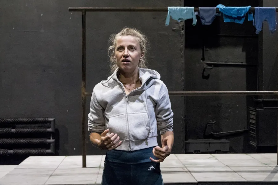 Zdjęcie przedstawia drugą próbę do spektaklu „Ifigenia ze Splott”. Agnieszka Skrzypczak znajduje się w centrum fotografii. Fotografia jest przybliżona, aktorkę widać od pasa w górę. Aktorka ma podniesioną głowę, patrzy ponad linię obiektywu. Ręce ma zgięte w łokciach. Palce ma lekko zgięte. Usta ma otwarte. Za aktorką widać fragment scenografii trzepaka. Na górnej poprzeczce trzepaka wisi błękitna odzież.