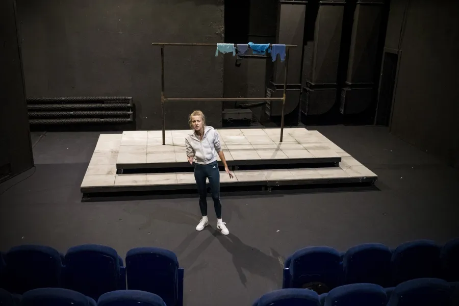 Zdjęcie przedstawia drugą próbę do spektaklu „Ifigenia ze Splott”. Agnieszka Skrzypczak znajduje się w centrum fotografii. Zdjęcie zostało wykonane od góry. Aktorka jest lekko przechylona w prawą stronę. Prawą rękę ma zgiętą w łokciu, dłonią wskazuje przed siebie. Lewą rękę ma wyprostowaną, lekko odchyloną od ciała. Patrzy przed siebie. Ma lekko otworzone usta. Przed aktorką widać dwa rzędy niebieskich foteli. Za aktorką widać fragment scenografii trzepaka. Na górnej poprzeczce trzepaka wisi błękitna odzież