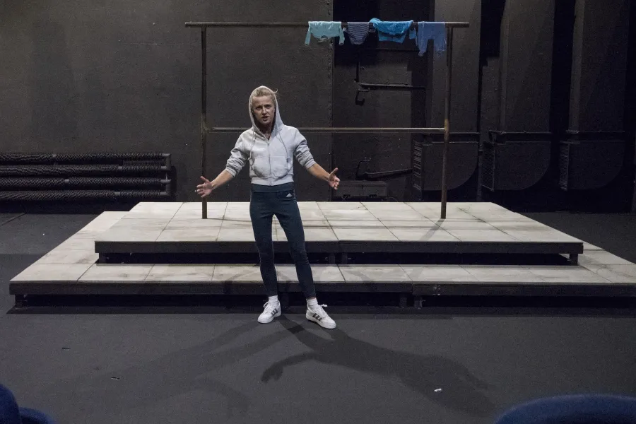 Zdjęcie przedstawia drugą próbę do spektaklu „Ifigenia ze Splott”. Przed podestem, na którym znajduje się scenografia trzepaka osiedlowego, stoi Agnieszka Skrzypczak. Ręce ma podniesione na wysokość bioder, rozłożone na boki. Jest lekko pochylona w prawo, lewą nogę ma lekko wysuniętą w bok. Patrzy się w obiektyw. Ma lekko otwarte usta. Na głowie ma założony kaptur białej bluzy. Na górnej poprzeczce trzepaka znajdującego się za nią, z prawej strony zawieszone są cztery sztuki błękitnej odzieży.