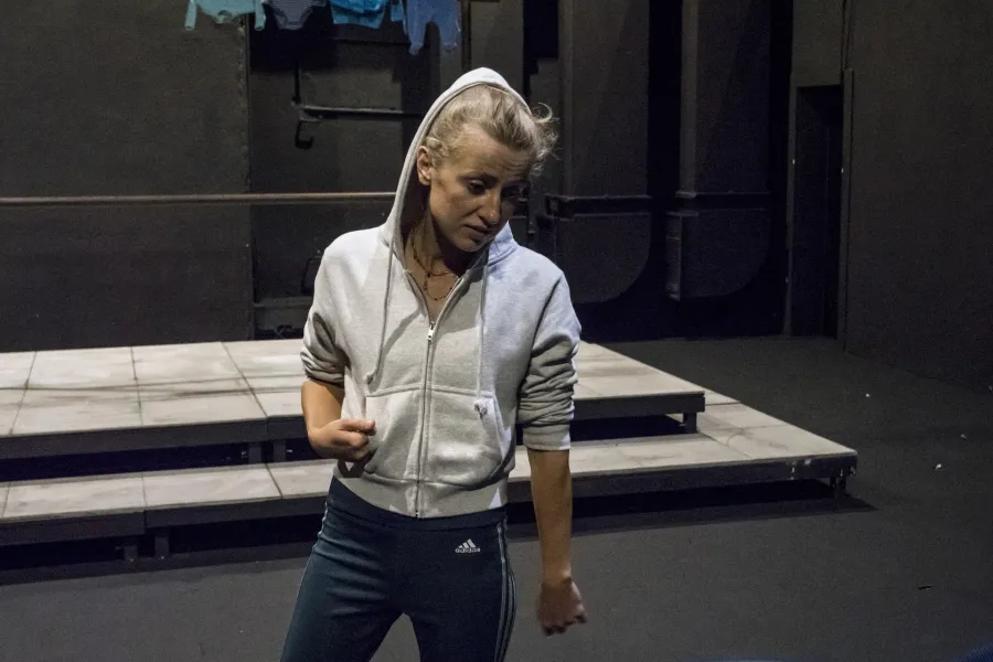 Zdjęcie przedstawia drugą próbę do spektaklu „Ifigenia ze Splott”. Agnieszka Skrzypczak stoi w centrum fotografii. Głowę ma lekko odwróconą w lewo, wzrok ma skierowany na podłogę. Prawą ręką trzyma kieszeń bluzy. Prawą nogę ma wysuniętą lekko w bok. Na głowie ma założony kaptur białej bluzy. Z lewej strony fotografii, za nią, stoi scenografia osiedlowego trzepaka na podeście. Na górnej poprzeczce trzepaka, z prawej strony zawieszone są cztery sztuki błękitnej odzieży.