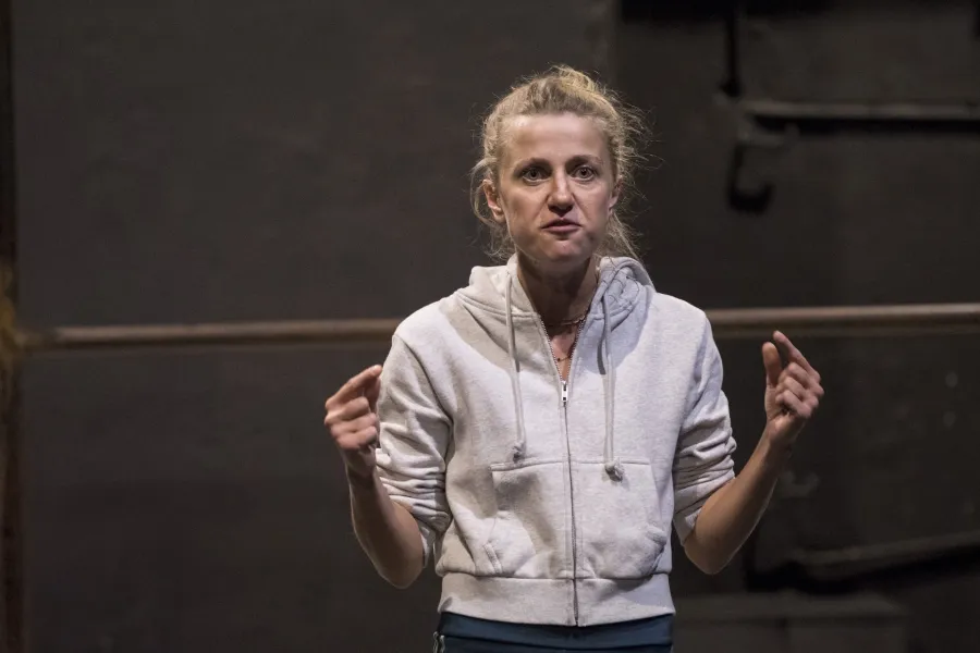 Zdjęcie przedstawia drugą próbę do spektaklu „Ifigenia ze Splott”. Agnieszka Skrzypczak stoi w centrum fotografii. Fotografia jest przybliżona, aktorkę widać od pasa w górę. Ręce ma podniesione na wysokość pasa. Aktorka wskazuje na siebie palcami wskazującymi obydwu rąk. Patrzy wprost, aczkolwiek nie w obiektyw. Za nią w tle za aktorką widać dolną poprzeczkę scenografii osiedlowego trzepaka.