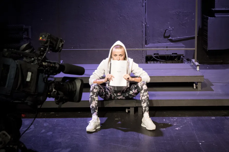 Zdjęcie przedstawia pierwszą próbę do spektaklu „Ifigenia ze Splott”. Na podeście w rozkroku siedzi Agnieszka Skrzypczak. Pod nosem trzyma scenariusz, przodem do obiektywu. Aktorka ubrana jest w ciemne legginsy w kwiaty, białe buty sportowe i białą bluzę, której kaptur ma założony na głowę. Uszy Agnieszki Skrzypczak ozdobione są dużymi kolczykami w kształcie okręgów w kolorze złota a włosy mocno zaczesane do tyłu. Za aktorką znajduje się scenografia w postaci trzepaka osiedlowego a przed nią kamera.  