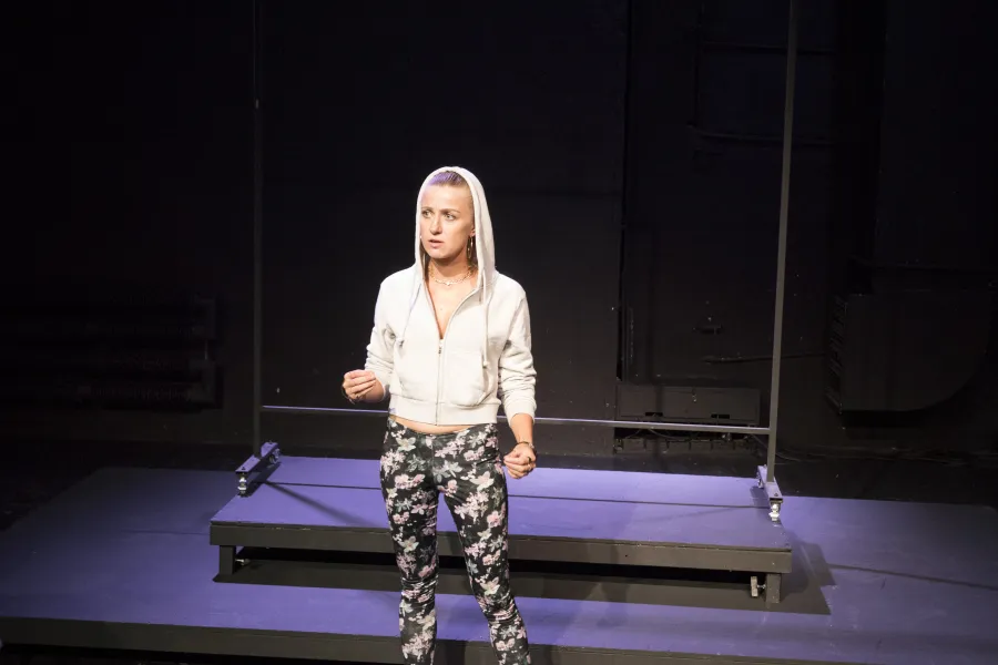 Zdjęcie przedstawia pierwszą próbę do spektaklu „Ifigenia ze Splott”. Przed podestami stoi Agnieszka Skrzypczak. Prawa ręka aktorki jest podniesiona na wysokość biodra a lewa na wysokość pępka. Wzrok Agnieszki Skrzypczak skierowany jest lekko do boku od obiektywu a jej usta delikatnie otwarte. Aktorka ubrana jest w ciemne legginsy w kwiaty, białe buty sportowe i białą bluzę, której kaptur ma założony na głowę. Za aktorką znajduje się scenografia w postaci trzepaka osiedlowego.