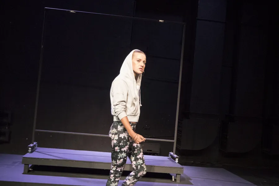 Zdjęcie przedstawia pierwszą próbę do spektaklu „Ifigenia ze Splott”. Przed podestami, bocznym profilem stoi Agnieszka Skrzypczak. Lewa noga aktorki jest zgięta w kolanie a prawa wyprostowana. Prawa ręka aktorki dotyka jej lewego uda a całą postawa jest lekko zgarbiona. Wzrok Agnieszki Skrzypczak sięga linii obiektywu, jej usta są otwarte a żuchwa lekko wysunięta do przodu. Aktorka ma założony kaptur na głowę a za nią znajduje się scenografia w postaci trzepaka osiedlowego.