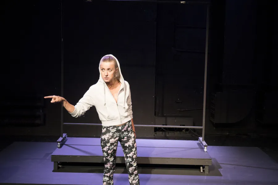 Zdjęcie przedstawia pierwszą próbę do spektaklu „Ifigenia ze Splott”. Przed podestami stoi Agnieszka Skrzypczak. Biodro aktorki jest lekko skręcone do boku, jej wzrok jest skierowany do boku. W tę samą stronę skierowany jest palec wskazujący prawej, zgiętej w łokciu ręki aktorki. Usta Agnieszki Skrzypczak są lekko otwarte a żuchwa wyciągnięta. Aktorka ma założony kaptur na głowę a za nią znajduje się scenografia w postaci trzepaka osiedlowego.