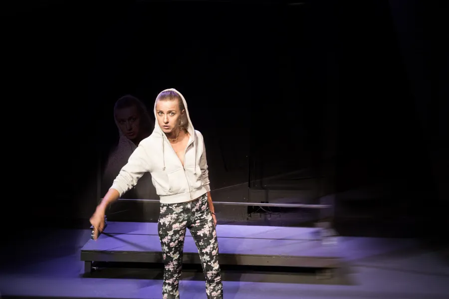 Zdjęcie przedstawia pierwszą próbę do spektaklu „Ifigenia ze Splott”. ”. Przed podestami stoi Agnieszka Skrzypczak. Biodro aktorki jest lekko skręcone do boku a wzrok jest skierowany w obiektyw. Palec wskazujący prawej ręki aktorki skierowany jest do dołu a usta są delikatnie otwarte. Aktorka ma założony kaptur na głowę a za nią znajduje się scenografia w postaci trzepaka osiedlowego.