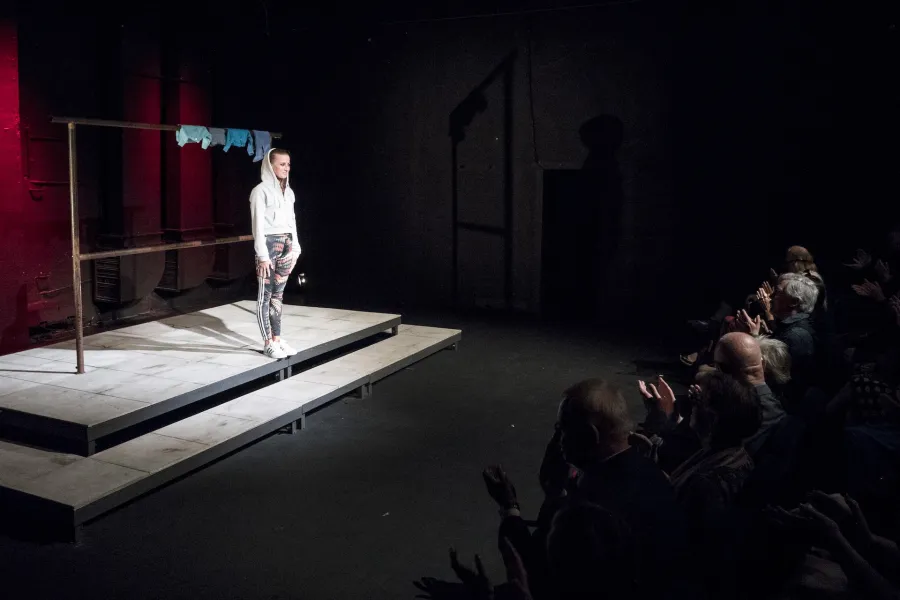 Zdjęcie przedstawia premierę spektaklu „Ifigenia ze Splott”. Agnieszka Skrzypczak znajduje się po lewej stronie fotografii. Jest odwrócona bokiem do obiektywu. Stoi wyprostowana, patrzy się na widownię przed sobą. Ma założony kaptur białej bluzy na głowie. Stoi na podeście, za nią widać scenografię trzepaka. Na górnej poprzeczce trzepaka wisi błękitna odzież. Aktorka i scena są oświetlone światłem reflektora. Po prawej stronie zdjęcia siedzi widownia, która patrzy na aktorkę i oklaskuje ją. 