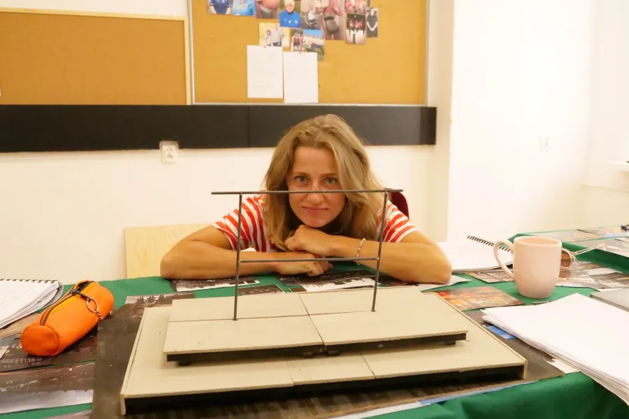 Zdjęcie przedstawia próbę stolikową do spektaklu „Ifigenia ze Splott”. Przy stole siedzi Agnieszka Skrzypczak z zgiętymi i rozłożonymi na stole łokciami. Głowa aktorki spoczywa na jej dłoniach blisko stołu a oczy skierowane są prosto w obiektyw. Przed aktorką na stole znajdują się fotografie i makieta osiedlowego trzepaka. Oprócz makiety po bokach, na stole znajduje się zeszyt, okulary, telefon, kubek, nożyczki, scenariusz i piórnik. Z tyłu wisi na ścianie tablica korkowa ze zdjęciami.  