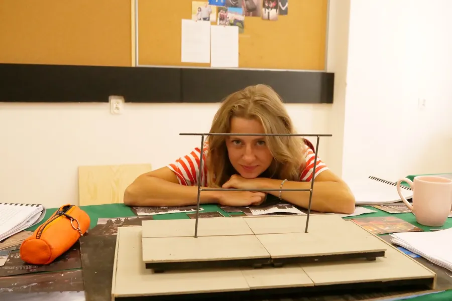 Zdjęcie przedstawia próbę stolikową do spektaklu „Ifigenia ze Splott”. Przy stole siedzi Agnieszka Skrzypczak z zgiętymi i rozłożonymi na stole łokciami. Głowa aktorki spoczywa na jej dłoniach blisko stołu a oczy skierowane są prosto w obiektyw. Przed aktorką na stole znajdują się fotografie i makieta osiedlowego trzepaka. Oprócz makiety po bokach, na stole znajduje się zeszyt, okulary, telefon, kubek, nożyczki, scenariusz i piórnik. Z tyłu wisi na ścianie tablica korkowa ze zdjęciami.  