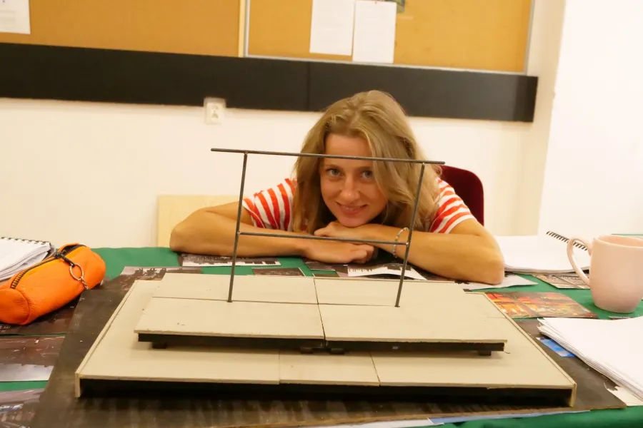 Zdjęcie przedstawia próbę stolikową do spektaklu „Ifigenia ze Splott”. Przy stole siedzi Agnieszka Skrzypczak z zgiętymi i rozłożonymi na stole łokciami. Głowa aktorki spoczywa na jej dłoniach blisko stołu a oczy skierowane są prosto w obiektyw. Na twarzy Agnieszki Skrzypczak widnieje delikatny uśmiech. Przed aktorką na stole znajdują się fotografie i makieta osiedlowego trzepaka. Oprócz makiety po bokach, na stole znajduje się zeszyt, okulary, telefon, kubek, scenariusz i piórnik. 