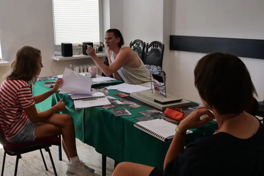 Zdjęcie przedstawia próbę stolikową do spektaklu „Ifigenia ze Splott”. Przy stole przodem siedzi reżyser Filip Gieldon a tyłem inspicjentka Marta Baraszkiewicz i Agnieszka Skrzypczak. Wzrok reżysera skierowany jest na aktorkę a jego lewa ręka jest zgięta w łokciu pod kątem prostym. Agnieszka Skrzypczak z lekko zgarbioną postawą przegląda scenariusz. Lewa ręka Marty Baraszkiewicz lekko dotyka jej brody. Na stole oprócz scenariuszy leży makieta trzepaka, fotografie, kubek i piórnik. 