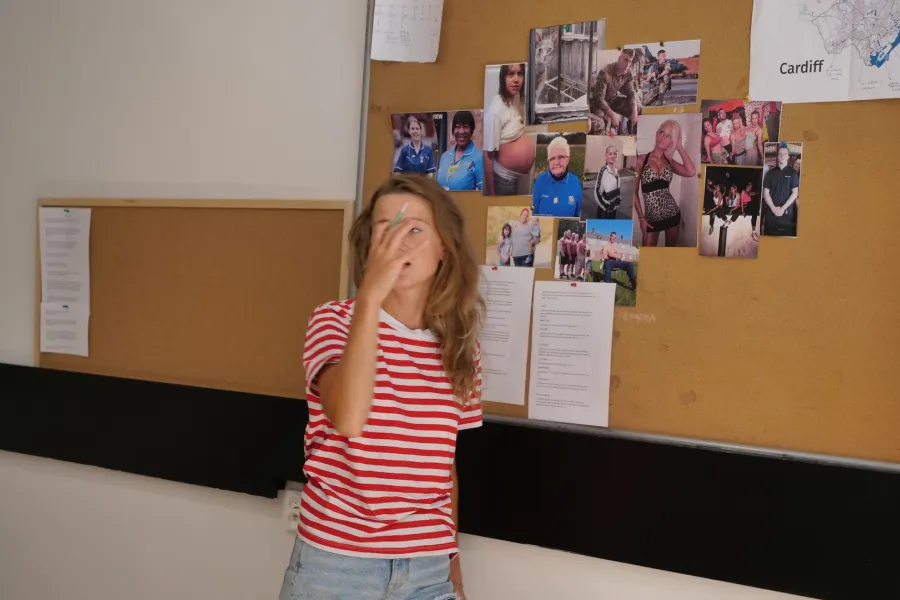 Zdjęcie przedstawia próbę stolikową do spektaklu „Ifigenia ze Splott”. Przy tablicy korkowej stoi Agnieszka Skrzypczak, której prawa ręka trzymająca długopis, zasłania jej prawie całą twarz. Na tablicy znajdują się fotografie różnych kobiet, mężczyzn i kota. Przypięta tam jest również mapa Cardiff oraz rożne teksty. 