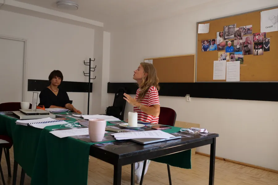 Zdjęcie przedstawia próbę stolikową do spektaklu „Ifigenia ze Splott”. Przy stole siedzi Agnieszka Skrzypczyk z inspicjentką Martą Baraszkiewicz. Głowa aktorki jest pochylona lekko do tyłu. Wzrok Marty Baraszkiewicz jest skierowany na Agnieszce Skrzypczyk. Na samym stole znajduje się scenariusz, kubki, klucze, zdjęcia, świeczka, słuchawki i telefony. Z tyłu na ścianie wisi tablica korkowa ze zdjęciami różnych osób. 