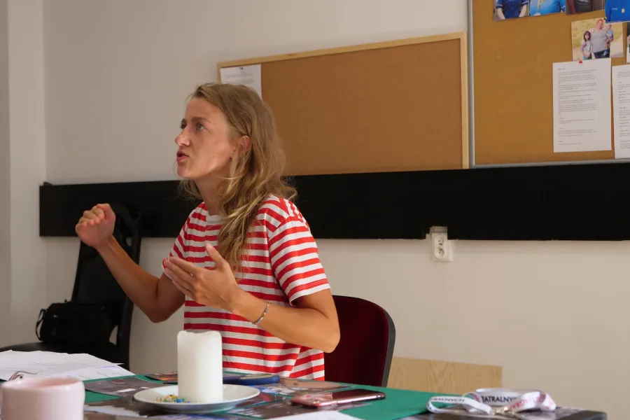 Zdjęcie przedstawia próbę stolikową do spektaklu „Ifigenia ze Splott”. Przy stole siedzi Agnieszka Skrzypczak ze zgiętymi, ale lekko wysuniętymi przed siebie rękami. Wzrok aktorki jest skierowany do boku, ponad obiektyw. Jej żuchwa jest wyciągnięta lekko do przodu a usta delikatnie otwarte. Przed aktorką na stole znajdują się fotografie, scenariusz, świeca, kubek i telefon. Z tyłu wisi na ścianie tablica korkowa ze zdjęciami.  