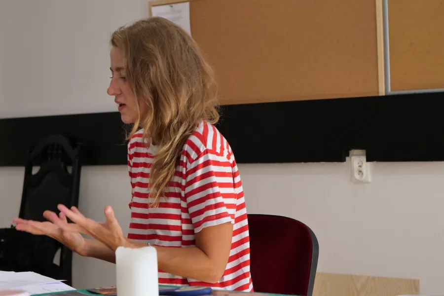 Zdjęcie przedstawia próbę stolikową do spektaklu „Ifigenia ze Splott”. Przy stole z wyciągniętymi przed siebie dłońmi siedzi Agnieszka Skrzypczyk. Wzrok aktorki skierowany jest w dół a jej usta są lekko otwarte. Na stole leży scenariusz a w tle widać tablice korkowe. 